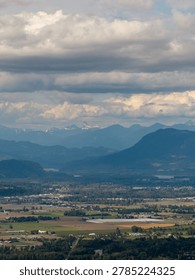 Dramatic layered clouds hover over Chilliwack valley farmland with distant snowcapped mountains in British Columbia, Canada.