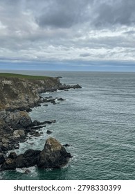 Dramatic layered rock cliffs stretching along the wild Cantabrian coastline, with scattered offshore rocks, turquoise green sea and a green clifftop meadow disappearing into the horizon under a stormy sky, Asturias, Spain