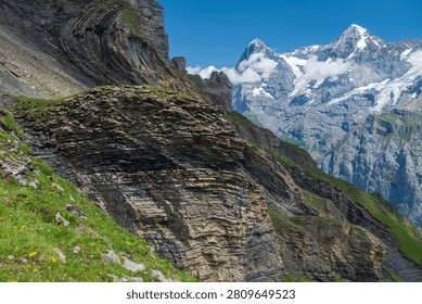 Dramatic layered rock face and green slope with snowcapped peaks under blue sky on a summer hike route above Lauterbrunnen Valley, Switzerland.
