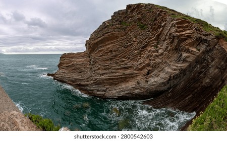 Dramatic layered rock formations meet the churning turquoise waters of the Cantabrian Sea under a cloudy sky
