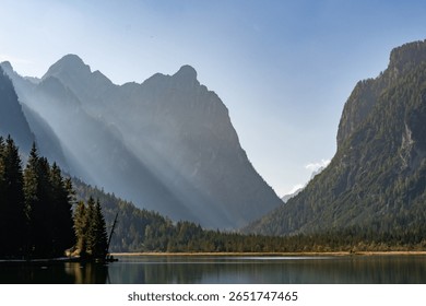 Dramatic light rays pierce through the majestic Dolomite mountains over Lago di Dobbiaco, Italy. A breathtaking alpine valley scene at dawn or dusk. Tranquil and inspiring.