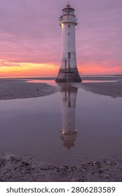 Dramatic lighthouse with perfect reflection on wet beach sand during golden hour sunset. Pink purple sky creates stunning seascape photography
