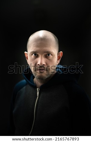 dramatic lighting short haired man serious look face portrait. unshaven looks intently holding attention. person dressed in black hoodie standing in a dark room. photo in the dark