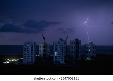 A dramatic lightning bolt illuminates the night sky above towering construction sites near the ocean. - Powered by Shutterstock