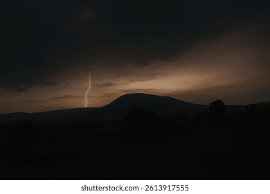 A dramatic lightning bolt strikes over a silhouetted mountain during dusk, creating a powerful contrast between the dark stormy clouds and the soft glow of the evening sky.