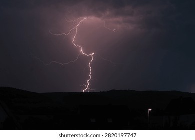 Dramatic Lightning Bolt Striking Over Rural Landscape During Night Thunderstorm

A powerful lightning bolt illuminates the dark night sky during an intense thunderstorm. The bright, jagged lightning strike branches dramatically across purple-tinted storm clouds, descending toward a silhouetted hillside landscape. Dark rooftops and a single streetlight are visible in the foreground, providing scale and context. Perfect for weather, natural phenomena, and dramatic atmosphere content.