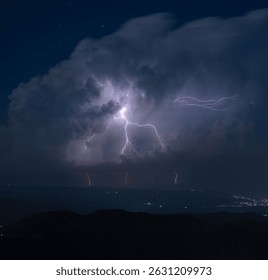 Dramatic lightning strikes burst through dense storm clouds over a dark mountainous terrain, revealing night lights in the distant cityscape.