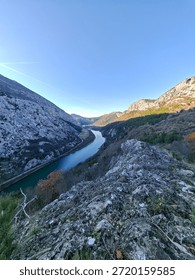 A dramatic limestone canyon opens beneath a rocky cliff, where a winding emerald river cuts through rugged mountains, sparse winter vegetation, and a wide, open sky.