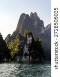 Dramatic limestone cliffs rising from a calm lake surrounded by tropical forest in Thailand under a clear blue sky.