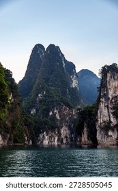 Dramatic limestone cliffs rising from a calm lake surrounded by tropical forest in Thailand under a clear blue sky.