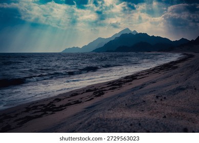 Dramatic long shot of a rugged beach in Sadah, Oman, featuring dark blue ocean waves crashing gently on the sandy shore. In the background, majestic mountains rise, silhouetted against a moody, cloudy sky with hints of sunlight breaking through. The overall atmosphere is serene, powerful, and mysterious. This evocative image is ideal for travel brochures, environmental campaigns, website backgrounds, editorial content, and advertising related to adventure, nature, or Middle Eastern tourism. It appeals to audiences interested in remote landscapes, natural beauty, and exotic destinations, offering a sense of calm and wonder.