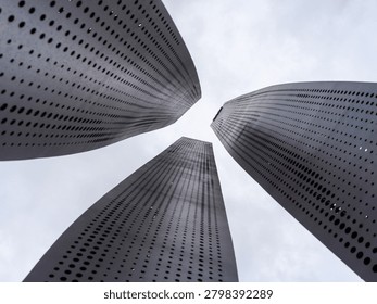 Dramatic look up view of three modern towering cylinders with black perforated dot pattern facades under a cloudy overcast sky. The photo represents futuristic industrial design, architectural engineering, urban abstract, and high tech structures.