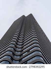 Dramatic low angle lookup view of a towering modern skyscraper with curved wavy glass facade rising high into cloudy overcast sky. The image portrays metropolitan growth, futuristic architecture, corporate dominance, real estate investment, and city landscape.