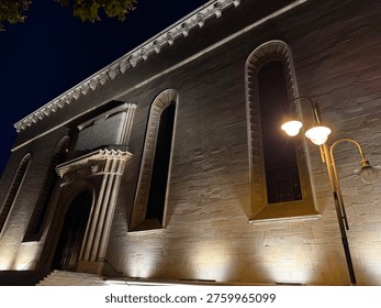 Dramatic low angle night view looking up at imposing historic stone ashlar building facade with ornate carved portal entrance columns decorative cornice arched windows with warm golden uplighting and vintage double street lamp with starburst light effect against deep blue night sky. Historic architecture concept photography in Eastern Europe.