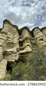 A dramatic low angle vertical shot looking up at magnificent, weathered volcanic tuff pillars featuring smooth, wavy caps in Cappadocia, Nevsehir, Turkey. The composition emphasizes the imposing height and layered geological textures of the cliffs, with a dark, hand-carved cave entrance visible at the base. Green bushes and wild vegetation frame the foreground under a dense, cloudy sky, creating a moody, mysterious, and awe-inspiring historic atmosphere.