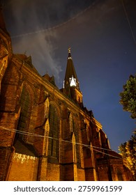 Dramatic low angle view looking up at imposing Gothic red brick church facade with pointed spire and golden cross illuminated against deep blue starry night sky with wispy clouds and aircraft contrail. Historic religious architecture concept photography in Poland.