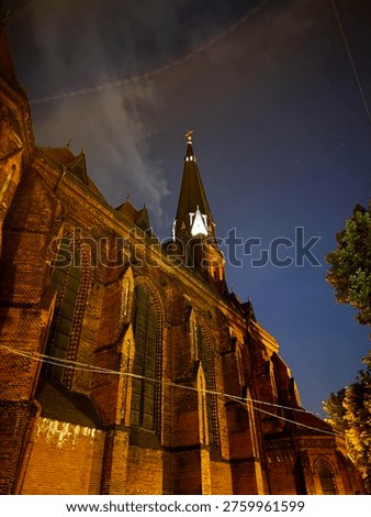Dramatic low angle view looking up at imposing Gothic red brick church facade with pointed spire and golden cross illuminated against deep blue starry night sky with wispy clouds and aircraft contrail. Historic religious architecture concept photography in Poland.