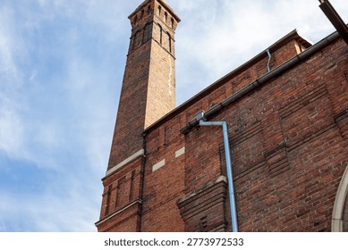 dramatic low angle view of a tall historic red brick tower rising above a matching brick building facade with decorative arched windows against a partly cloudy blue sky