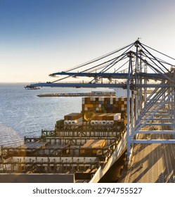 A dramatic low-angle aerial view showcasing towering port cranes positioned above a massive container ship at a busy maritime terminal. The scene highlights global trade, logistics, cargo transportation, and modern shipping infrastructure, emphasizing the scale and efficiency of international supply chain operations.