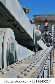Dramatic low-angle close-up of the structural steel beams, riveted construction, suspension elements, and illuminated ornamental lamp posts of a historic bridge. The composition emphasizes perspective, industrial craftsmanship, and nineteenth-century civil engineering with the iconic bridge gateway framed in the background under a clear blue summer sky.