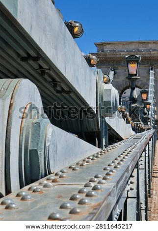 Dramatic low-angle close-up of the structural steel beams, riveted construction, suspension elements, and illuminated ornamental lamp posts of a historic bridge. The composition emphasizes perspective, industrial craftsmanship, and nineteenth-century civil engineering with the iconic bridge gateway framed in the background under a clear blue summer sky.