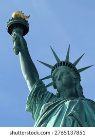 A dramatic low-angle close-up view of the Statue of Liberty shot from below, looking up at the iconic copper figure against a clear bright blue sky. The composition highlights the raised arm holding the golden torch, the radiating crown with seven spikes, and the finely detailed facial expression of Lady Liberty in serene, dignified repose. The verdigris green patina of the oxidized copper contrasts beautifully with the warm gold of the torch flame and the clean azure sky, creating a powerful and visually striking image. This perspective emphasizes the monumental scale and enduring grandeur of one of the world's most recognized symbols of freedom, democracy, and the American spirit. Located on Liberty Island in New York Harbor, the statue was a gift from France to the United States and has welcomed millions of immigrants and visitors since 1886. Ideal for travel and tourism content, American history and culture editorials, patriotic campaigns, Independence Day visuals, immigration and freedom concepts, New York City destination marketing, and architectural landmark photography projects.