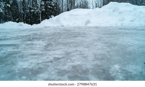 A dramatic low-angle ground-level photograph looking across a thick, textured sheet of frozen glare ice toward large plowed snowbanks and winter forest trees on a cold, overcast day.