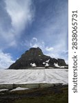 A dramatic, low-angle landscape view capturing the imposing north face of Clements Mountain towering over a massive summer snowfield at Logan Pass in Glacier National Park. The foreground features a vast, textured expanse of melting white snow blanketed across the steep alpine tundra slope. In the background, the colossal, stratified dark rock peak rises sharply towards a dynamic sky filled with wispy, sweeping cirrus clouds and deep blue accents. Distinct bands of seasonal snow remain tucked neatly into the rugged crevices and lower ledges of the mountain, highlighting the wild, high-altitude terrain of the Montana Rockies.