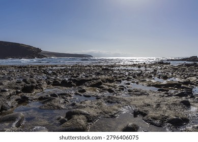 A dramatic low-angle perspective emphasizes the rich texture of wet volcanic pebbles and rocks reflecting the brilliant midday sun. The blinding silver glint on the ocean surface creates a powerful, high-contrast atmosphere of raw coastal beauty.
