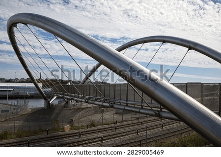 A dramatic low-angle perspective looking up through the sleek stainless steel tubular arches and suspension cables of the Celtic Gateway footbridge in Holyhead, Anglesey, Wales. The bold architectural curve of the pedestrian bridge spans over railway tracks leading towards the port, set against a wide blue sky filled with wispy cirrus clouds.