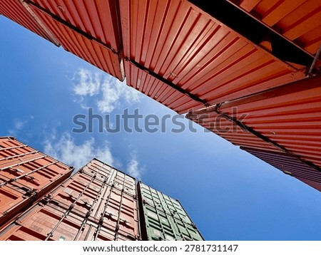 Dramatic low-angle perspective of stacked red and green cargo shipping containers rising toward a bright blue sky with clouds. Industrial maritime logistics and global transportation infrastructure at a commercial shipping terminal