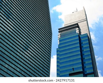 A dramatic low-angle perspective of two modern glass skyscrapers set against a bright blue sky with soft white clouds. On the left, a towering building with a curved facade is covered in a dense pattern of horizontal glass slats that reflect the surrounding light. To the right, a multi-tiered building stands tall, showcasing a darker blue glass exterior and a distinct grey concrete section at the top equipped with various antennas.