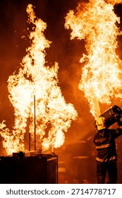 A dramatic, low-angle photograph captures a firefighter in full protective gear—including a helmet and a dark jacket with reflective stripes—operating a hose. The firefighter is directing a powerful stream of water or fire-suppressing foam into a colossal blaze. The massive flames are bright orange and yellow, dominating the upper portion of the frame against a pitch-black background. A thick cloud of warm-hued smoke and countless sparkling droplets of water or ash are visible, creating a highly intense and hazardous environment. The scene effectively conveys the danger and the immediate, courageous action of emergency services.