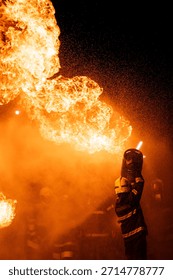 A dramatic, low-angle photograph captures a firefighter in full protective gear—including a helmet and a dark jacket with reflective stripes—operating a hose. The firefighter is directing a powerful stream of water or fire-suppressing foam into a colossal blaze. The massive flames are bright orange and yellow, dominating the upper portion of the frame against a pitch-black background. A thick cloud of warm-hued smoke and countless sparkling droplets of water or ash are visible, creating a highly intense and hazardous environment. The scene effectively conveys the danger and the immediate, courageous action of emergency services.