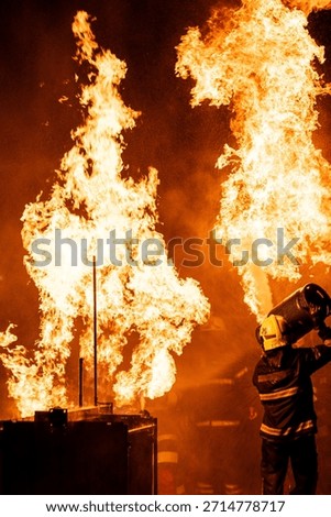 A dramatic, low-angle photograph captures a firefighter in full protective gear—including a helmet and a dark jacket with reflective stripes—operating a hose. The firefighter is directing a powerful stream of water or fire-suppressing foam into a colossal blaze. The massive flames are bright orange and yellow, dominating the upper portion of the frame against a pitch-black background. A thick cloud of warm-hued smoke and countless sparkling droplets of water or ash are visible, creating a highly intense and hazardous environment. The scene effectively conveys the danger and the immediate, courageous action of emergency services.