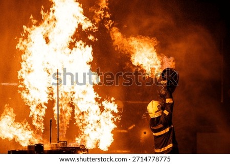 A dramatic, low-angle photograph captures a firefighter in full protective gear—including a helmet and a dark jacket with reflective stripes—operating a hose. The firefighter is directing a powerful stream of water or fire-suppressing foam into a colossal blaze. The massive flames are bright orange and yellow, dominating the upper portion of the frame against a pitch-black background. A thick cloud of warm-hued smoke and countless sparkling droplets of water or ash are visible, creating a highly intense and hazardous environment. The scene effectively conveys the danger and the immediate, courageous action of emergency services.