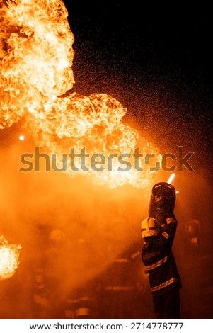A dramatic, low-angle photograph captures a firefighter in full protective gear—including a helmet and a dark jacket with reflective stripes—operating a hose. The firefighter is directing a powerful stream of water or fire-suppressing foam into a colossal blaze. The massive flames are bright orange and yellow, dominating the upper portion of the frame against a pitch-black background. A thick cloud of warm-hued smoke and countless sparkling droplets of water or ash are visible, creating a highly intense and hazardous environment. The scene effectively conveys the danger and the immediate, courageous action of emergency services.