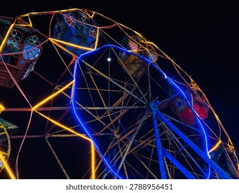 A dramatic low-angle photograph capturing the elaborate network of lights on a vintage, weathered Ferris wheel at night. The intricate metal frame, showing signs of age and rust, is brilliantly traced with vibrant lines of neon-style LED lighting, forming a complex, abstract web of geometric patterns. Bold lines of warm golden-yellow and electric blue lights intersect, creating a series of overlapping stars and polygons (like hexagons and pentagons). Several traditional, decorative passenger gondolas, each in a different color like red, blue, and gold with detailed geometric patterns, are visible in the upper sections of the frame. A small, bright crescent moon is clearly visible against the inky black sky through a gap in the structure. The aged texture of the blue-painted metal contrasts with the piercing clarity of the modern lights. The dynamic perspective makes the structure feel both grand and intricate, set against the profound darkness of the night.