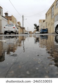 A dramatic low-angle photograph capturing a rain-soaked street in a generic urban setting. The foreground features a large puddle reflecting blurred cars and buildings, with a defocused cityscape and 