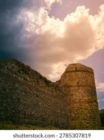 A dramatic low-angle shot of an ancient stone fortress wall and circular tower under a dynamic sky filled with contrasting dark storm clouds and bright, warm sunlight.