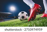 A dramatic low-angle shot captures a vibrant red soccer cleat poised above a classic black and white ball on lush green grass, illuminated by a bright stadium floodlight at night.