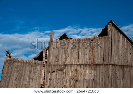 A dramatic, low-angle shot of the deteriorating wooden side of an old barn, with its roofline collapsing against a clear blue sky with white clouds.