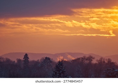 A dramatic low-angle shot of a glowing orange and yellow sunset sky with heavy clouds hanging over a dark, snow-dusted forest and distant mountain peaks.