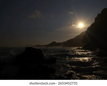Dramatic low-angle shot of a glowing sunset over the ocean, silhouetting rugged rocks and distant hills. Sun rays pierce the sky, creating a powerful and mystical coastal scene.