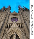 A dramatic low-angle shot of a grand Gothic cathedral facade, featuring twin spires, a large rose window, and an arched entrance, under a blue evening sky.