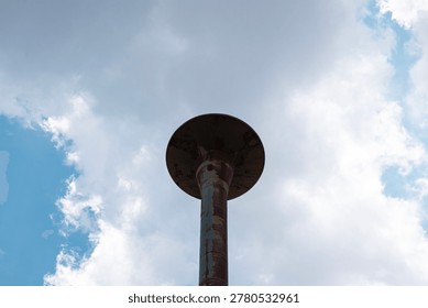 Dramatic low-angle shot of a heavily rusty, derelict flared-top standpipe water tower soaring against a partly cloudy blue sky in the countryside. The discolored, textured metal surface is sharply focused.
