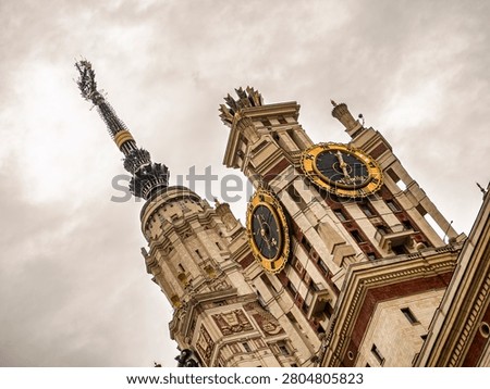 A dramatic low-angle shot of the iconic Moscow State University main building featuring golden clock faces and a towering spire against a cloudy sky, showcasing grand Stalinist architecture.