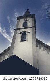 Dramatic low-angle shot of a tall white church bell tower with a sharp spire reaching towards a deep blue sky with streaky white clouds.