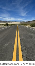 A dramatic low-angle symmetrical view stretches along Nevada's Highway 50, known as the Loneliest Road in America, with bold double yellow center lines converging toward pale sand dunes and rugged mountains under a vivid blue sky. The vast Great Basin desert landscape flanks both sides of the empty two-lane highway, with sparse sagebrush and arid terrain extending to the horizon. Ideal for travel, road trip, adventure, and American West editorial or commercial content.