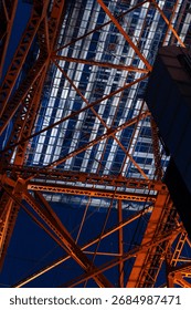 A dramatic low-angle vertical shot of Tokyo Tower’s illuminated orange steel framework, showcasing dense lattice patterns against a deep night sky, symbolizing strength, scale, and urban architecture.